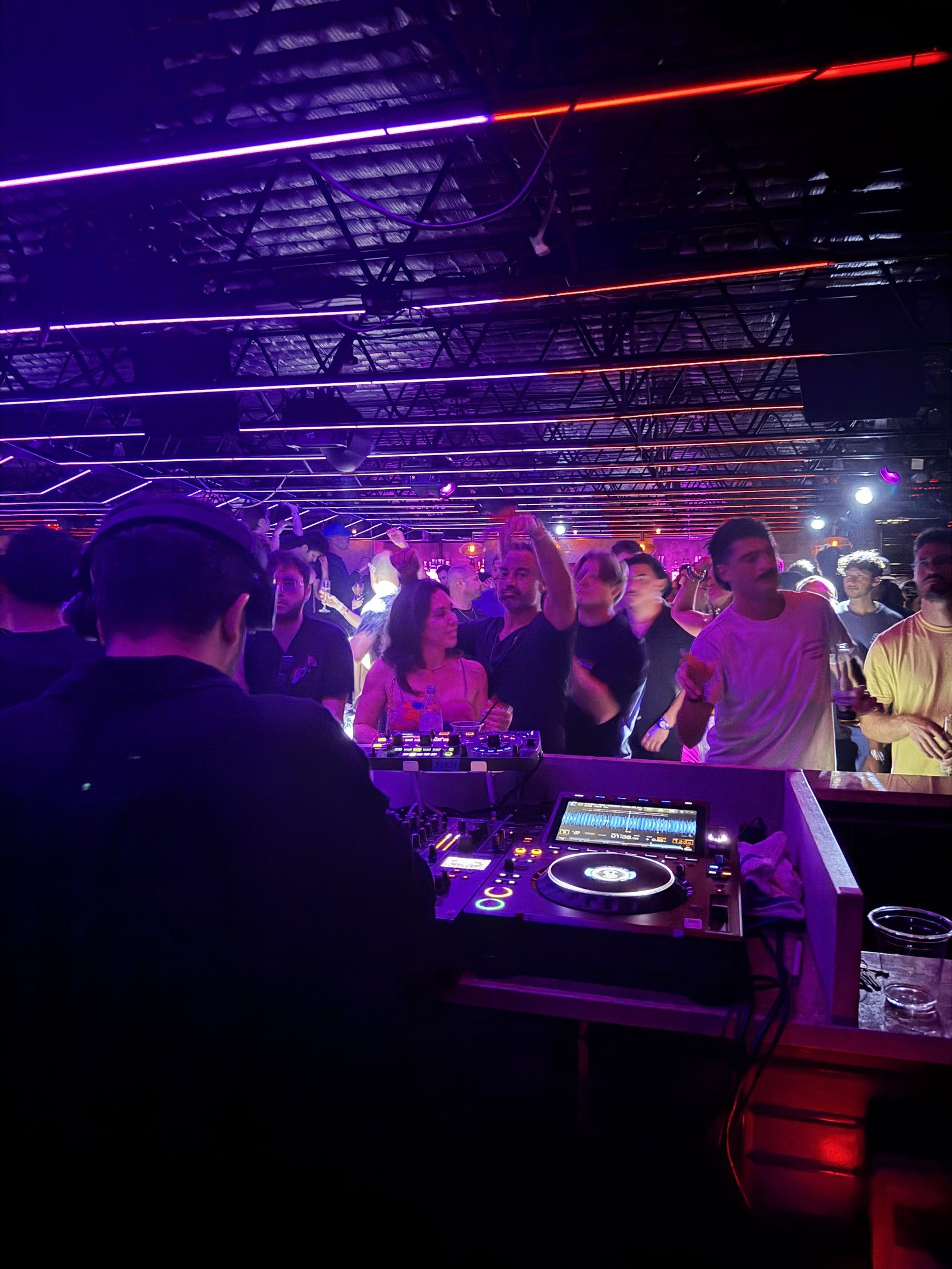 A packed dance floor under purple lights seen from behind the DJ booth